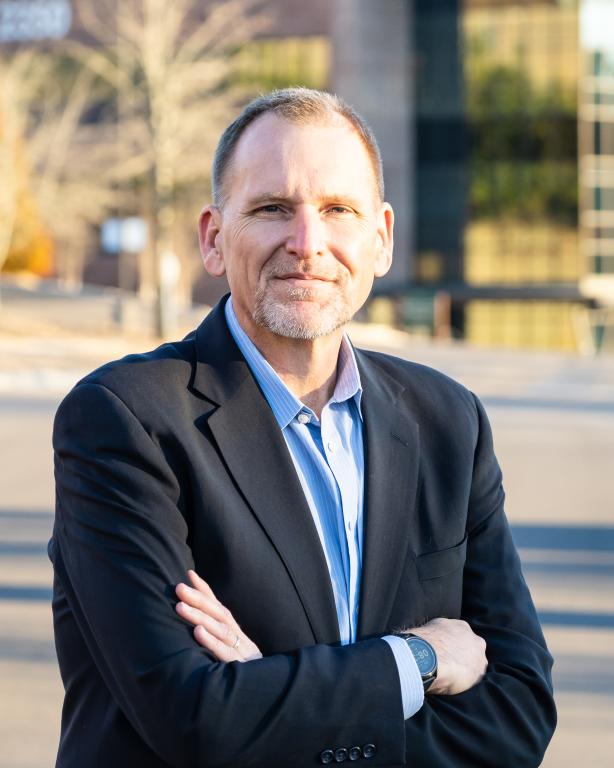 Photo of a man in a black jacket suit with a blue shirt crossing his arms outside. He is smiling.