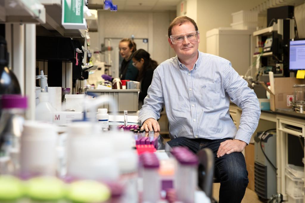 Hugh O'Neil, director or ORNL's Center for Structural Molecular Biology is sitting in the lab on a stool, hand on desk with glasses on. There are lab related items blurred in the foreground.