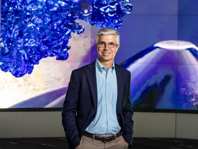 Shaun gleason is standing in front of a blue and purple screen posing for a photo with his hands in his pocket